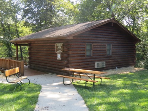 A rustic log cabin surrounded by trees, featuring a porch, picnic tables, and a pathway leading to the entrance.
