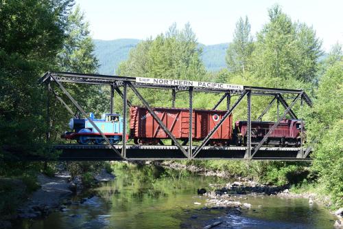 A blue train engine and red freight car cross a metal bridge over a stream, surrounded by trees and mountains.