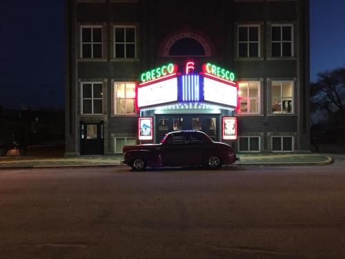 A vintage car parked in front of a brightly lit theater marquee at dusk.
