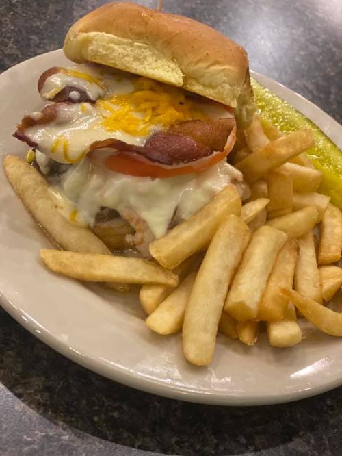 A plate with a cheeseburger topped with bacon, tomato, and cheese, served with a side of golden French fries and a pickle.
