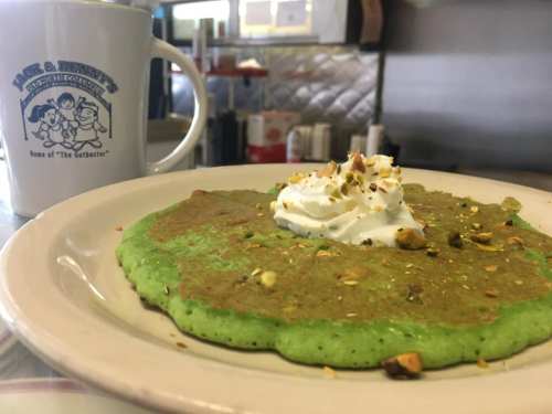 A green pancake topped with whipped cream and nuts, next to a coffee mug with a cartoon logo.