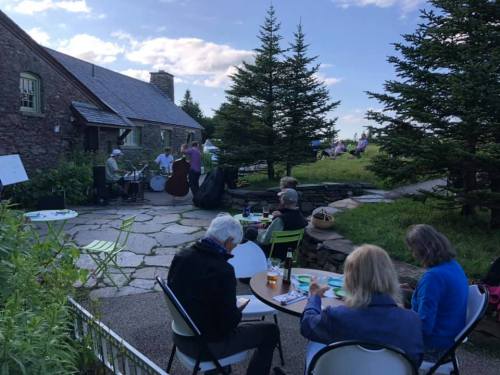 A group enjoys music outdoors at a scenic venue, with people seated at tables and greenery surrounding them.