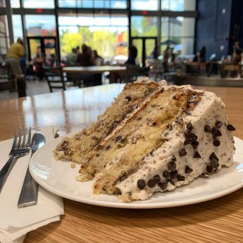 A slice of layered cake with chocolate chips and frosting on a white plate, set on a wooden table in a café.