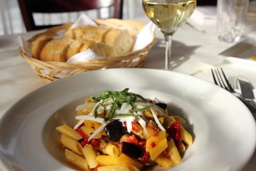A plate of pasta with vegetables, topped with basil, alongside a glass of white wine and a basket of bread.