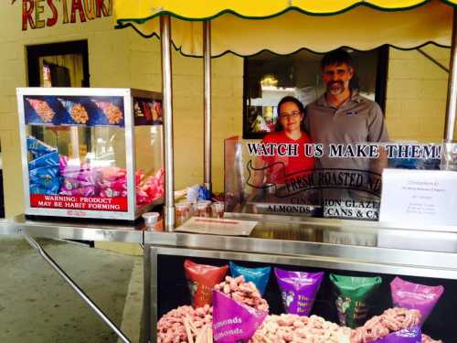 A man and woman stand behind a snack stand with colorful bags of roasted nuts and a sign that reads "WATCH US MAKE THEM!"