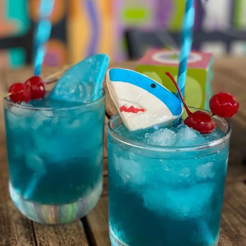 Two vibrant blue cocktails with ice, garnished with cherries and a shark-shaped decoration, served in clear glasses.