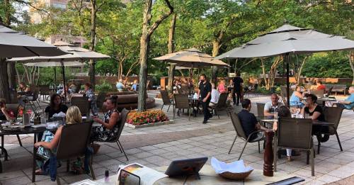 Outdoor dining area with patrons seated under umbrellas, surrounded by greenery and flowers, enjoying a lively atmosphere.