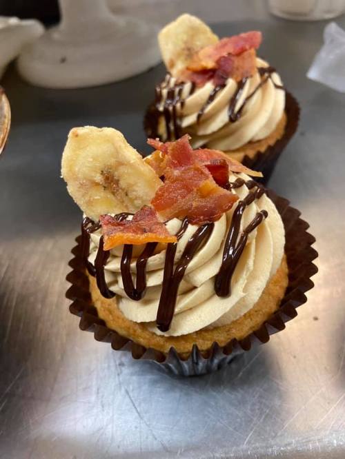 Two cupcakes topped with swirls of frosting, chocolate drizzle, banana chips, and crispy bacon bits.