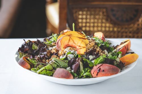 A vibrant salad with mixed greens, grilled peaches, walnuts, and crumbled cheese in a white bowl on a table.