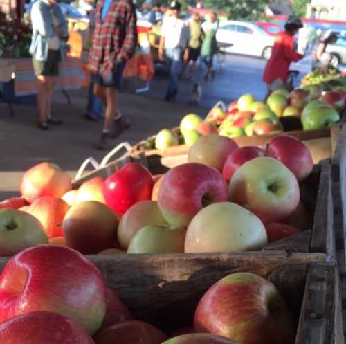 Fresh apples in wooden crates at a bustling farmers market with people browsing in the background.