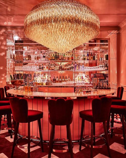 A stylish bar with a large chandelier, illuminated shelves of liquor, and red velvet seating.