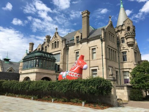 A large inflatable Santa sits in front of a grand, historic mansion under a blue sky with scattered clouds.
