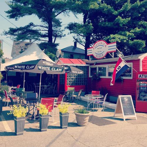 A colorful diner with outdoor seating, umbrellas, and potted plants, surrounded by trees on a sunny day.