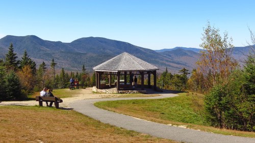 A scenic view of a gazebo on a hill, surrounded by trees and mountains under a clear blue sky.