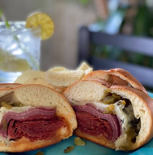 A close-up of a sliced sandwich filled with layers of meat, cheese, and pickles, served with potato chips and a drink.