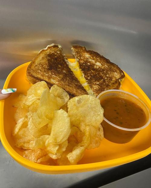 A yellow plate with a grilled cheese sandwich, crispy potato chips, and a small cup of dipping sauce.