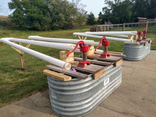 Two metal troughs with red hand pumps and white pipes, set on wooden supports in a grassy area.