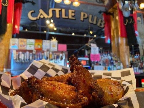 A plate of chicken wings in front of a bar with "Castle Pub" sign and various drinks in the background.