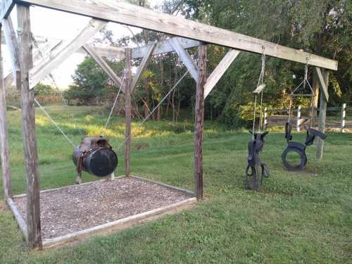 A wooden swing set with a barrel swing and tire swing, set in a grassy area with trees in the background.