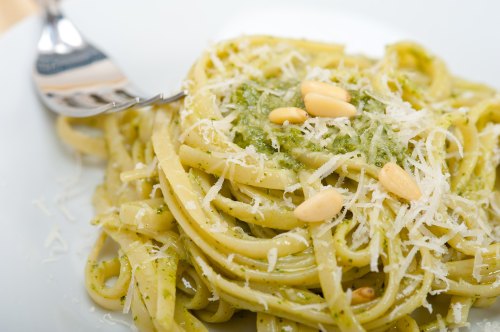 A plate of pasta with pesto sauce, topped with pine nuts and grated cheese, served with a fork on the side.