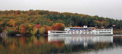 A white boat docked on a calm lake, surrounded by vibrant autumn foliage reflecting in the water.