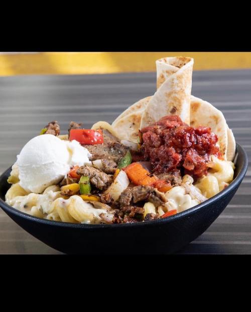 A bowl of macaroni topped with grilled meat, vegetables, salsa, and a dollop of sour cream, with tortillas on the side.