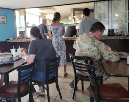 A casual dining scene with patrons at a counter, including a person in military uniform and others seated at tables.