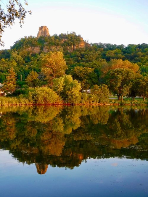 A serene landscape featuring a rocky hill reflected in a calm lake, surrounded by lush trees and greenery.