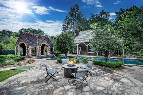 A serene outdoor space featuring a stone patio, fire pit, pool, and charming structures surrounded by trees.