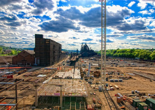 A construction site with cranes, steel structures, and industrial buildings under a cloudy sky.