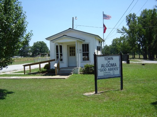 A small white building with a sign reading "Town of Algoma 'God Abides'" and a flag, surrounded by green grass.