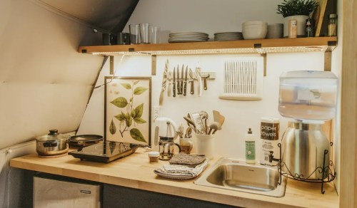 A cozy kitchen with wooden shelves, utensils, a sink, and a water dispenser, featuring plants and decorative items.