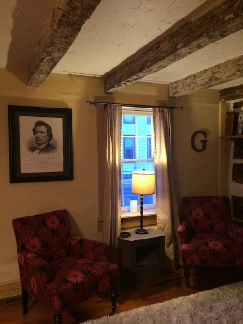 Cozy room with two patterned chairs, a lamp, and a window, featuring a portrait on the wall and rustic wooden beams.