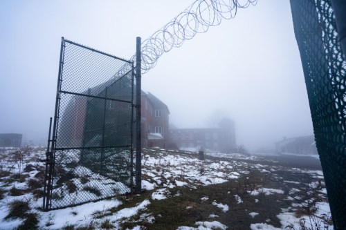 Foggy scene of an abandoned building behind a chain-link fence, with barbed wire and patches of snow on the ground.