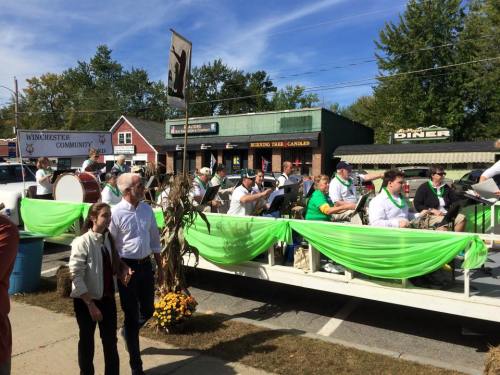 A parade float with musicians in green attire, surrounded by festive decorations and local shops in the background.