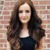 A young woman with long, wavy brown hair poses in front of a brick wall, wearing a black top.
