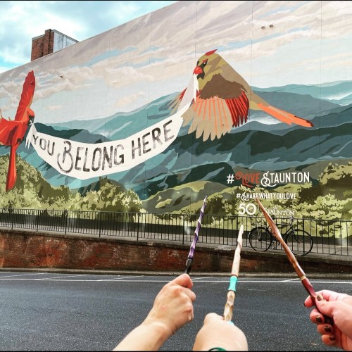 A colorful mural featuring birds and the text "You Belong Here" with people holding wands in the foreground.