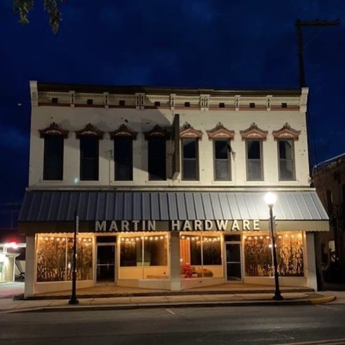 Historic building at night with "MARTIN HARDWARE" sign, featuring decorative windows and warm exterior lighting.
