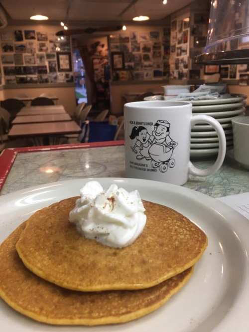 A plate of pancakes topped with whipped cream, with a coffee mug and a cozy diner setting in the background.