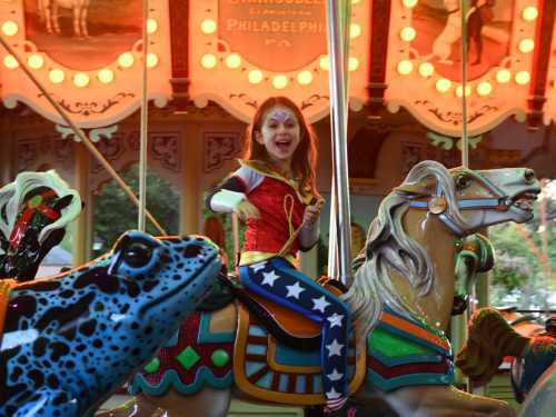 A joyful girl dressed as Wonder Woman rides a colorful carousel horse, surrounded by vibrant lights and decorations.