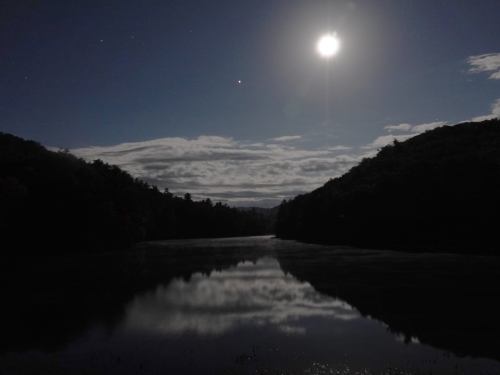 A serene night scene with a bright moon reflecting on a calm river, surrounded by dark hills and a starry sky.