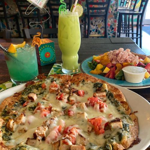 A table with a spinach chicken pizza, a colorful fruit salad, and two vibrant cocktails in a lively restaurant setting.