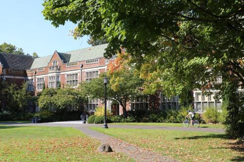 A sunny campus scene featuring a brick building surrounded by trees and a path, with a person riding a bicycle.