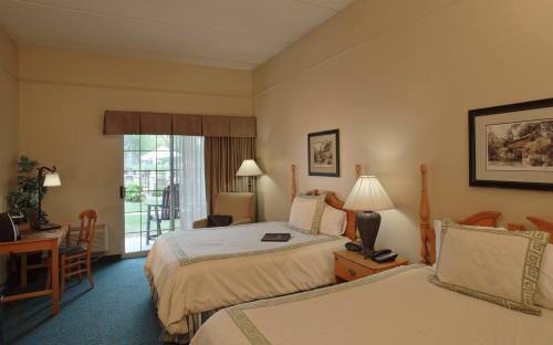 A cozy hotel room with two beds, a desk, and a sliding door leading to a patio area. Soft lighting and warm decor.