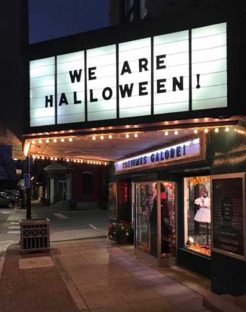 A theater marquee at night reads "WE ARE HALLOWEEN!" with a costume shop visible in the window.