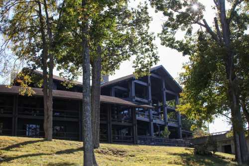 A large, dark wooden building surrounded by trees, set on a grassy slope under a clear blue sky.