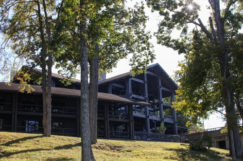 A large, dark wooden building surrounded by trees, set on a grassy slope under a clear blue sky.