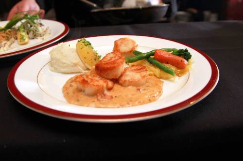 A plate of seared scallops on a creamy sauce, served with mashed potatoes and colorful sautéed vegetables.