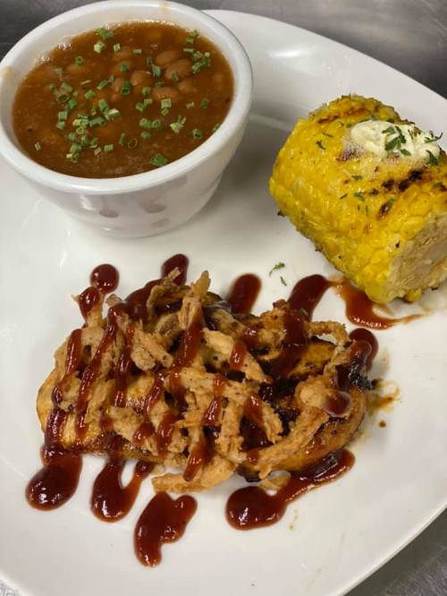 A plate featuring grilled chicken with BBQ sauce, corn on the cob, and a bowl of brown beans, garnished with chives.