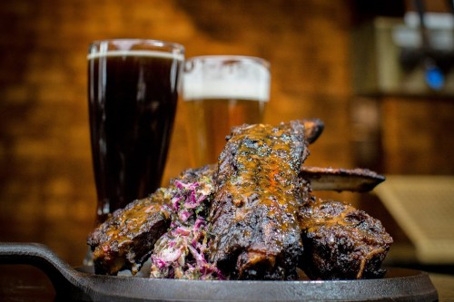 A plate of glazed ribs with coleslaw, accompanied by two glasses of dark and light beer.